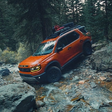 2027 Honda Passport TrailSport Elite in Sunset Orange profile driving through a rocky stream in the forest (Mid-Size SUV).